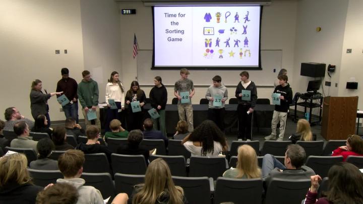 A line of students in front of an auditorium of people playing a college admissions game with a projection displayed behind them. 