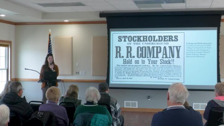 Cate Waldecker from the Detroit Historical Society, left of frame, talking to a crowd with a slide projected next to her. 