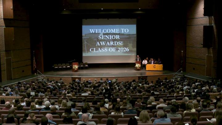A screen with the text Welcome to Senior Wards Class of 2026 with a full auditorium of people. 