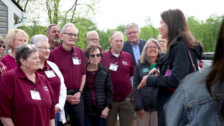 A group of people, left of frame, listening to a women speak, right of frame. 