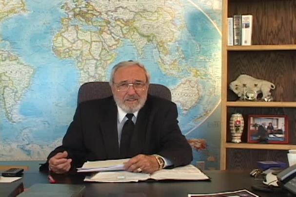 A man sitting center with a map behind him and a bookcase to the right. 