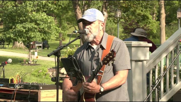 A man sings into a microphone outside holding a guitar. 