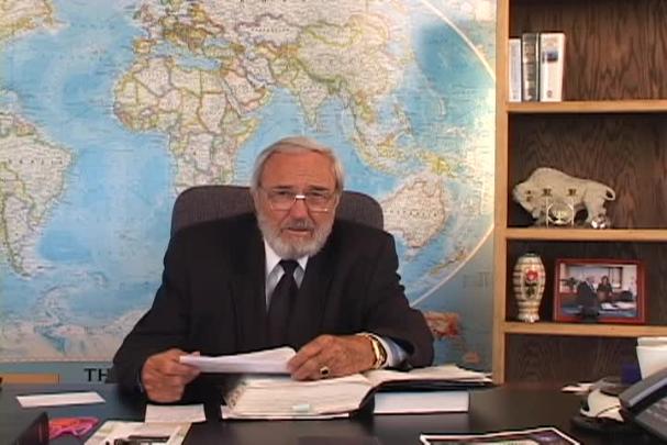 A man sits at a desk with a map behind him. 