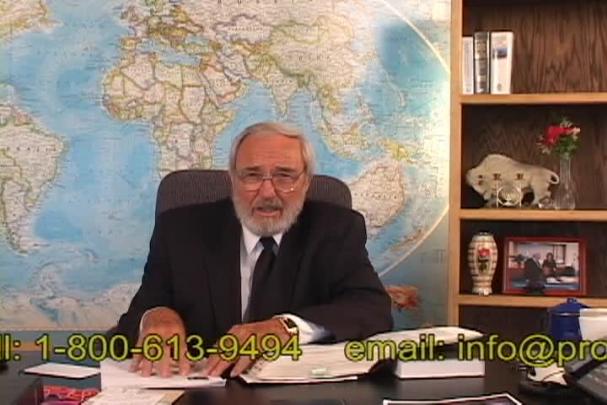 A man sitting center with a map behind him and a bookcase to the right. 