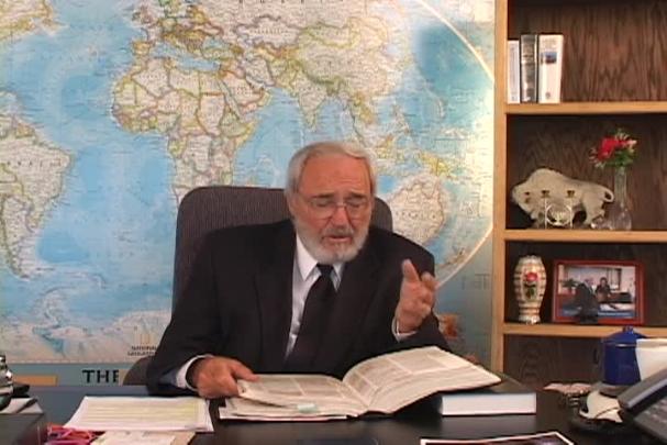 A man sitting center motioning with his left hand with a map behind him and a bookcase to the right. 