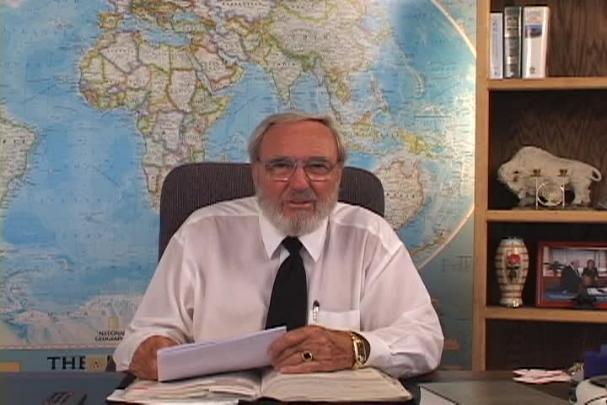 A man sitting center with a map behind him and a bookcase to the right. 