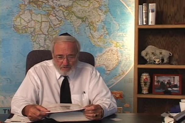 A man sitting center with a map behind him and a bookcase to the right. 