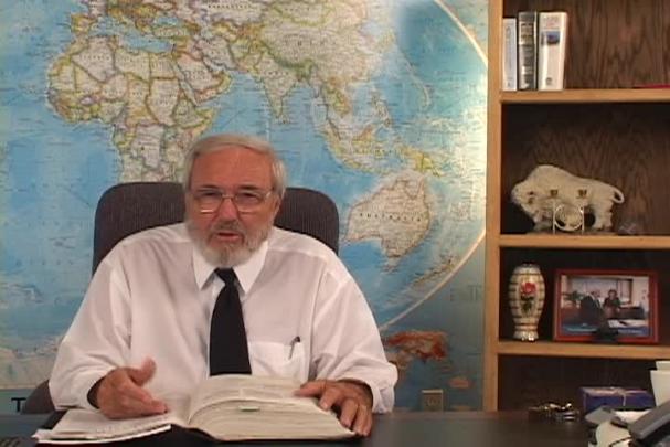 A man sitting center with a map behind him and a bookcase to the right. 