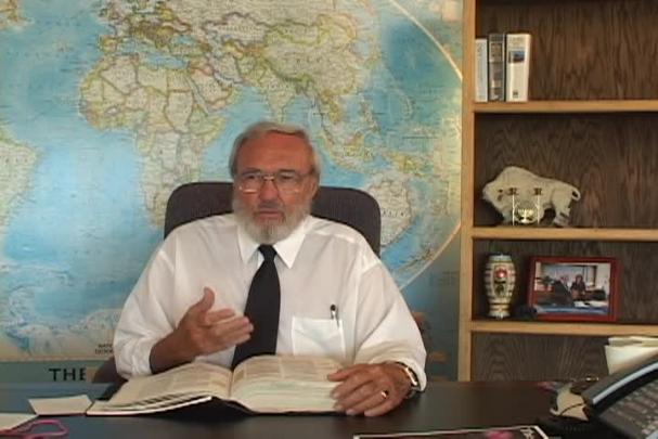 A man sitting center with a map behind him and a bookcase to the right. 
