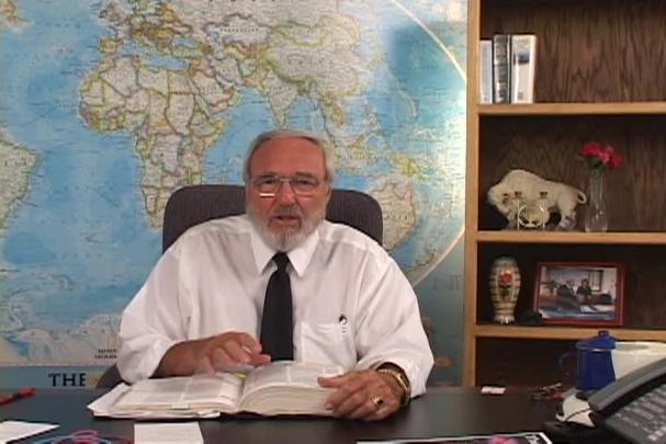 A man sitting center with a map behind him and a bookcase to the right. 