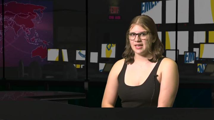 A woman sits at a desk with a virtual set behind her. 